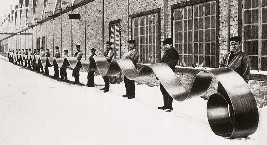 Historical photo of a line of about 20 men holding a long strip steel outside a factory building.