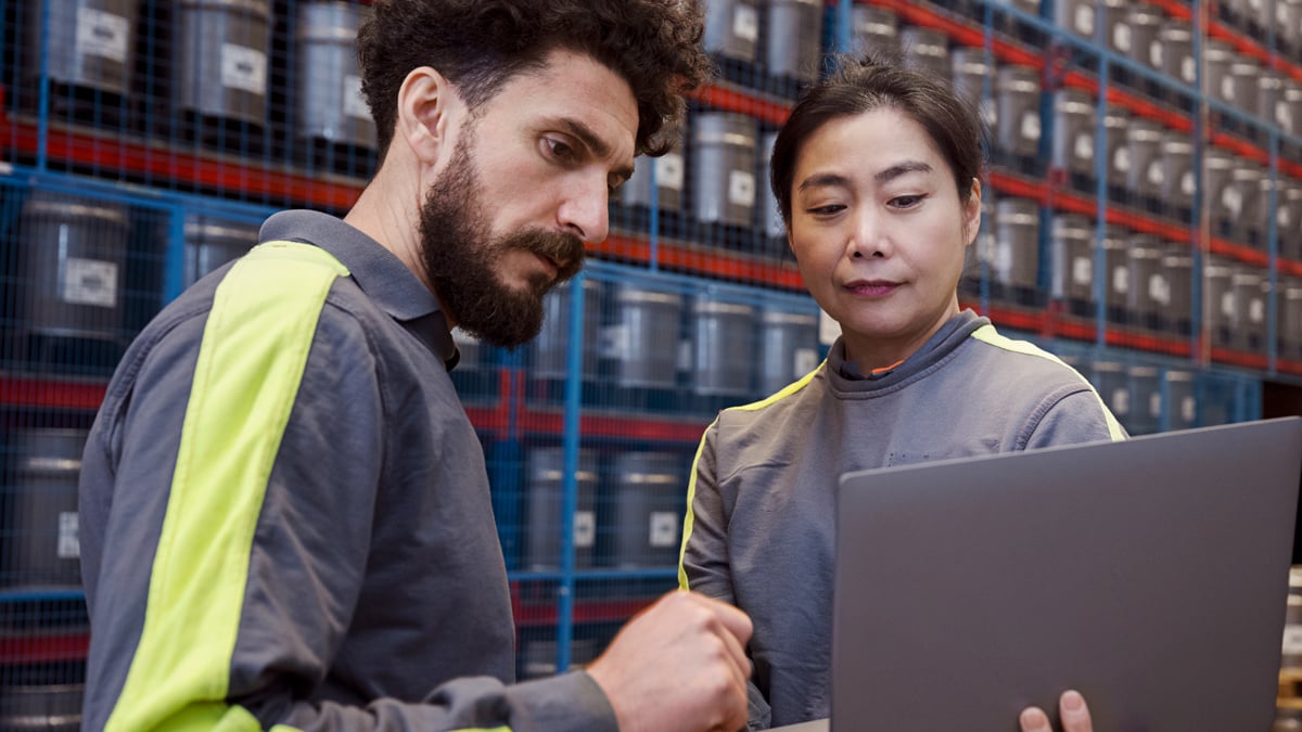 A woman and a man standing between two warehouse shelfs. The woman is holding a laptop and both are looking at it.