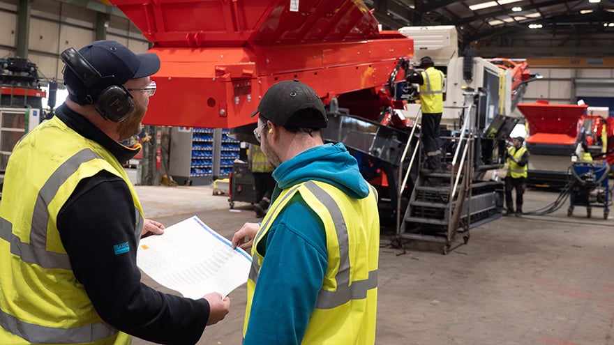 Two men in safety clothes in a production environment for heavy equipment. One of them has a drawing in his hand which both are looking at.